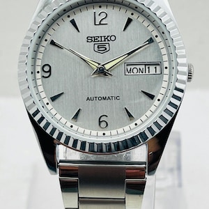 May include: Silver-toned Seiko 5 automatic wristwatch with a fluted bezel. The watch face is silver with black markers and hands, displaying the day and date. The watch band is a matching silver metal link style.