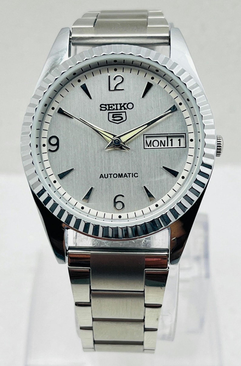 May include: Silver-toned Seiko 5 automatic wristwatch with a fluted bezel. The watch face is silver with black markers and hands, displaying the day and date. The watch band is a matching silver metal link style.