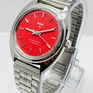 May include: A vintage wristwatch with a bright red face and silver-toned metal band. The watch face features the text "hmt" and "PARA SHOCK 17 JEWELS". The watch is displayed on a clear plastic stand.