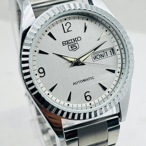 May include: Silver-toned Seiko 5 automatic watch with a silver dial, black markers, and a day-date display. The watch has a fluted bezel and a stainless steel band. The watch face displays the text "SEIKO 5 AUTOMATIC".