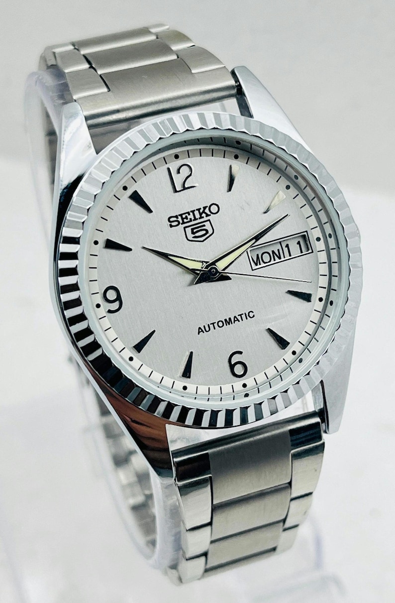 May include: Silver-toned Seiko 5 automatic watch with a silver dial, black markers, and a day-date display. The watch has a fluted bezel and a stainless steel band. The watch face displays the text "SEIKO 5 AUTOMATIC".