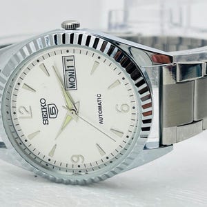May include: A silver-toned Seiko 5 automatic watch with a white face. The watch features a day and date display, luminous hands, and a stainless steel band. The watch has a fluted bezel and is designed for everyday wear.