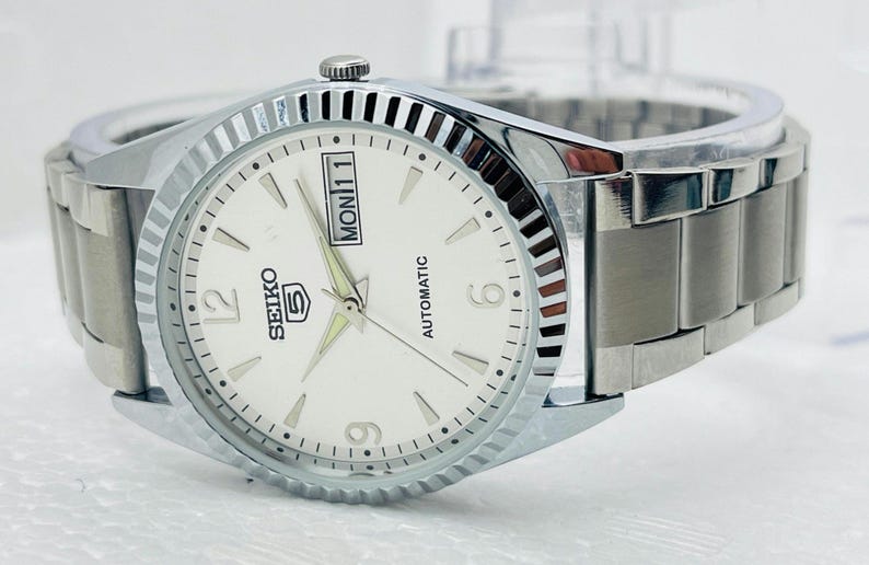 May include: A silver-toned Seiko 5 automatic watch with a white face. The watch features a day and date display, luminous hands, and a stainless steel band. The watch has a fluted bezel and is designed for everyday wear.