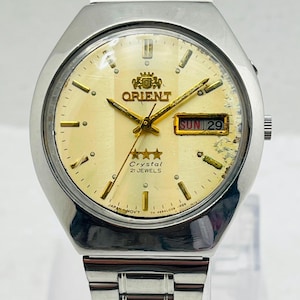 May include: A silver-tone stainless steel Orient wristwatch with a pale yellow dial, gold-colored hands and hour markers, and a day-date window displaying SUN 29. The dial features the Orient logo, three stars, and the text Crystal 21 JEWELS.