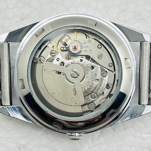 May include: Close-up of a silver-toned wristwatch with a transparent back, revealing the intricate mechanical movement. The watch has a silver band and a detailed internal mechanism with gears and springs.