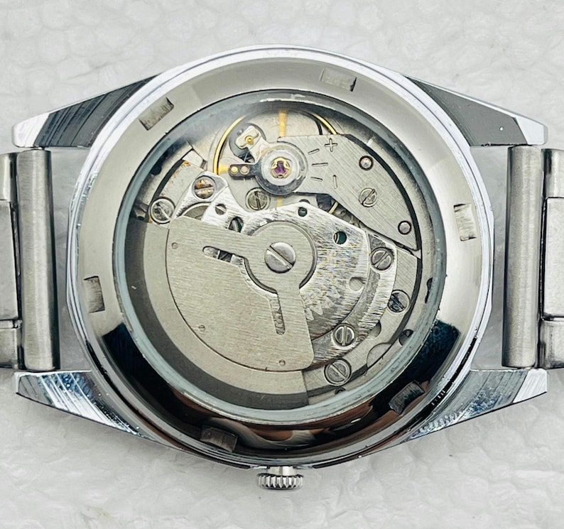 May include: Close-up of a silver-toned wristwatch with a transparent back, revealing the intricate mechanical movement. The watch has a silver band and a detailed internal mechanism with gears and springs.