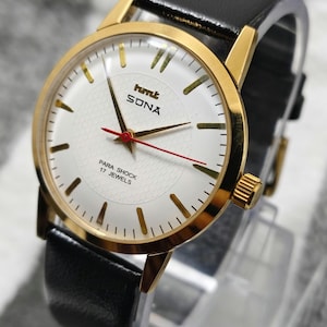 May include: A gold-plated wristwatch with a white face and black leather strap. The watch face displays the text "hmt SONA" and "PARA SHOCK 17 JEWELS". The watch has gold hands and markers, with a red second hand.
