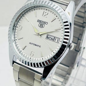 May include: Silver-toned Seiko 5 automatic watch with a white dial. The watch features a fluted bezel, a stainless steel band, and a day-date display. The watch face displays the day as "MON 11".