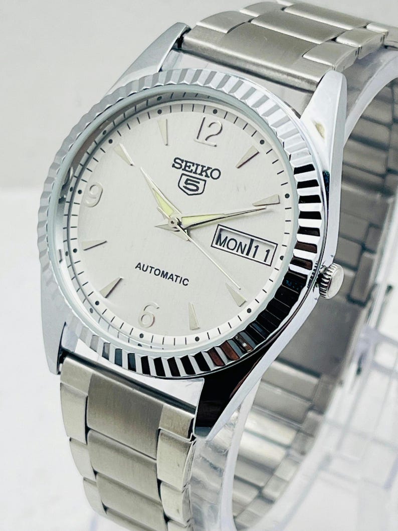 May include: Silver-toned Seiko 5 automatic watch with a white dial. The watch features a fluted bezel, a stainless steel band, and a day-date display. The watch face displays the day as "MON 11".
