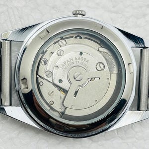 May include: Close-up of a silver-toned wristwatch with a transparent back, revealing the intricate internal mechanism. The watch face displays the text "JAPAN 6309A SEVENTEEN 17 JEWELS". The watch has a silver band and a textured bezel.