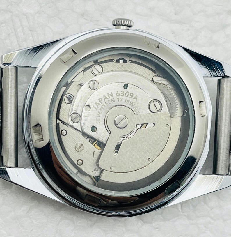 May include: Close-up of a silver-toned wristwatch with a transparent back, revealing the intricate internal mechanism. The watch face displays the text "JAPAN 6309A SEVENTEEN 17 JEWELS". The watch has a silver band and a textured bezel.