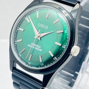 Peut inclure: Une montre-bracelet noire avec un cadran vert. Le cadran affiche la marque "ORIS" et le texte "17 JEWELS SHOCK PROOF". La montre a des aiguilles et des marqueurs argentés, et un bracelet en métal noir.