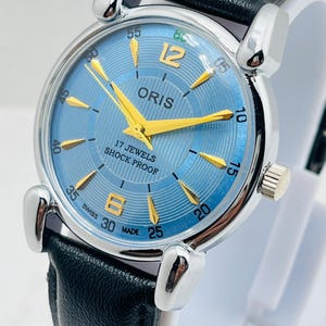 Op de afbeelding: Een vintage Oris polshorloge met een lichtblauwe wijzerplaat, gouden wijzers en markeringen en een zwarte leren band. De wijzerplaat toont de tekst "ORIS", "17 JEWELS" en "SHOCK PROOF". Het horloge heeft een roestvrijstalen kast.