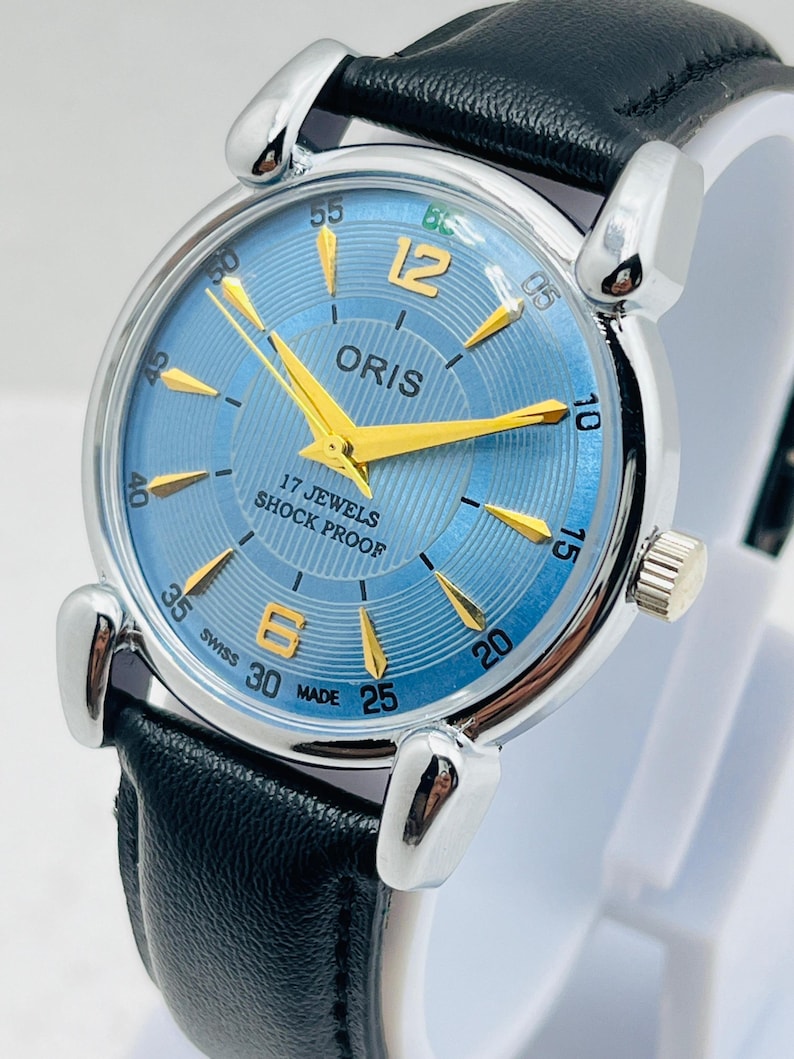 Op de afbeelding: Een vintage Oris polshorloge met een lichtblauwe wijzerplaat, gouden wijzers en markeringen en een zwarte leren band. De wijzerplaat toont de tekst "ORIS", "17 JEWELS" en "SHOCK PROOF". Het horloge heeft een roestvrijstalen kast.