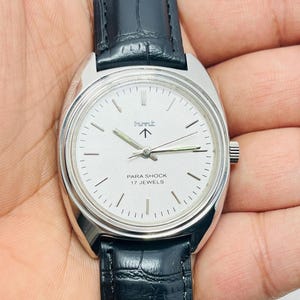 May include: A silver-toned wristwatch with a white face and black leather band. The watch face features the text "PARA SHOCK 17 JEWELS" and the brand logo. The watch is held in a person's hand.