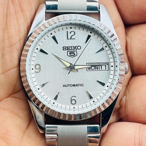 May include: Silver-toned Seiko 5 automatic watch with a white dial, featuring black hour markers, and a day-date display. The watch has a fluted bezel and a stainless steel band. The watch face displays the word "AUTOMATIC".