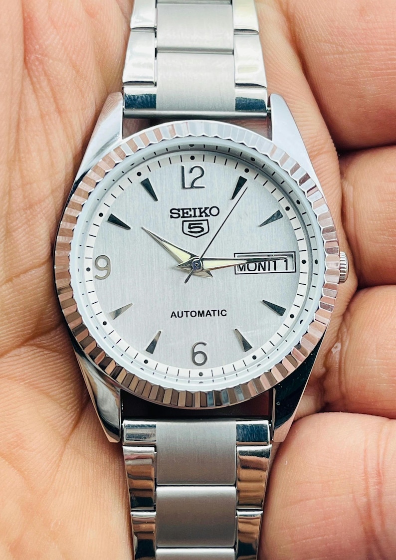 May include: Silver-toned Seiko 5 automatic watch with a white dial, featuring black hour markers, and a day-date display. The watch has a fluted bezel and a stainless steel band. The watch face displays the word "AUTOMATIC".