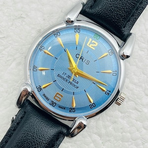 Op de afbeelding: Een polshorloge met een lichtblauwe wijzerplaat en goudkleurige wijzers en markeringen. Het horloge heeft een zilverkleurige kast en een zwarte leren band. De wijzerplaat heeft de tekst "CRIS", "17 JEWELS" en "SHOCK PROOF".