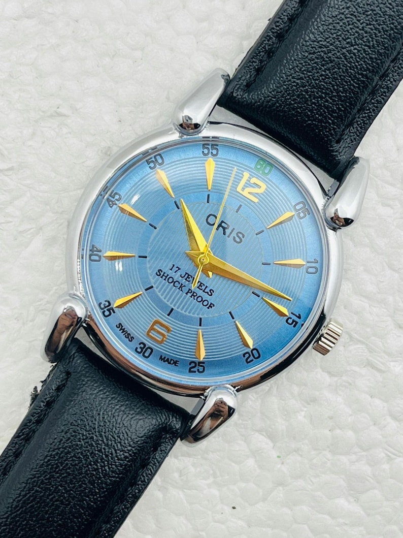 Op de afbeelding: Een polshorloge met een lichtblauwe wijzerplaat en goudkleurige wijzers en markeringen. Het horloge heeft een zilverkleurige kast en een zwarte leren band. De wijzerplaat heeft de tekst "CRIS", "17 JEWELS" en "SHOCK PROOF".