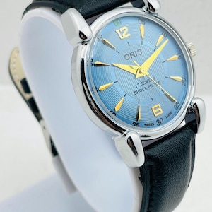 Op de afbeelding: Een zilverkleurig polshorloge met een lichtblauwe wijzerplaat en gouden wijzers. Het horloge heeft een zwarte leren band. Op de wijzerplaat staat het woord "ORIS", en de woorden "17 JEWELS" en "SHOCK PROOF".