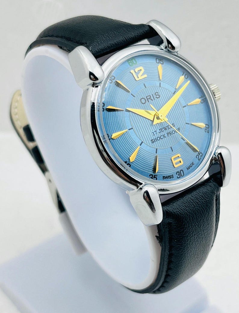 Op de afbeelding: Een zilverkleurig polshorloge met een lichtblauwe wijzerplaat en gouden wijzers. Het horloge heeft een zwarte leren band. Op de wijzerplaat staat het woord "ORIS", en de woorden "17 JEWELS" en "SHOCK PROOF".