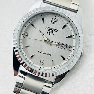 May include: A silver Seiko 5 automatic wristwatch with a fluted bezel and a silver dial. The watch features black hour markers, luminous hands, and a day-date display. The stainless steel bracelet is visible.
