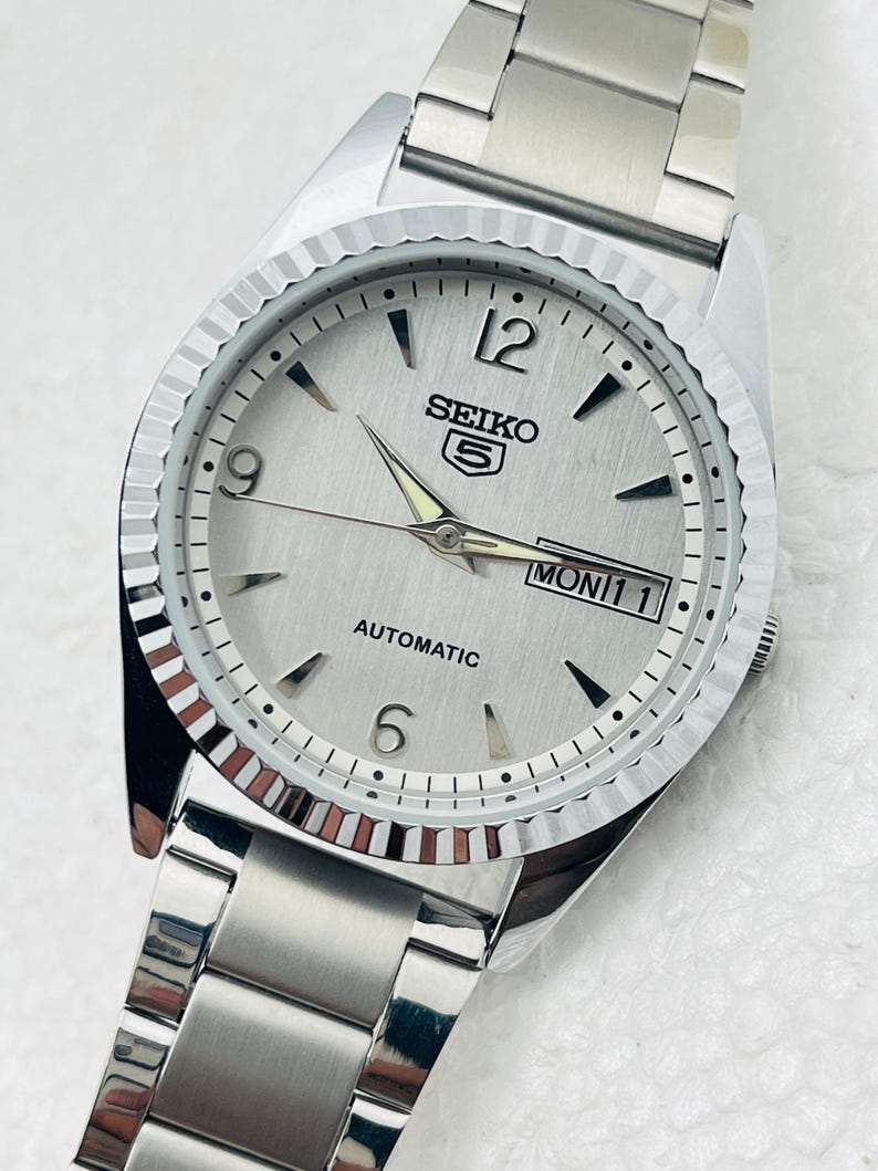 May include: A silver Seiko 5 automatic wristwatch with a fluted bezel and a silver dial. The watch features black hour markers, luminous hands, and a day-date display. The stainless steel bracelet is visible.
