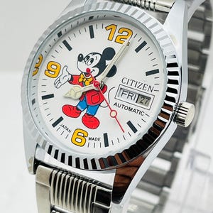 Op de afbeelding: Een zilveren Citizen automatisch polshorloge met een witte wijzerplaat met Mickey Mouse. Het horloge heeft een datumweergave en een zilveren band. De cijfers 6, 9 en 12 zijn geel. Het horloge is gemarkeerd met "Japan Made".