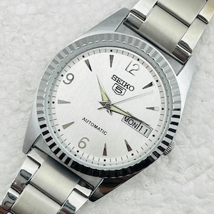 May include: Silver-toned Seiko 5 automatic watch with a white dial. The watch features black hour markers, a day-date display, and a fluted bezel. The watch band is made of silver-toned metal.