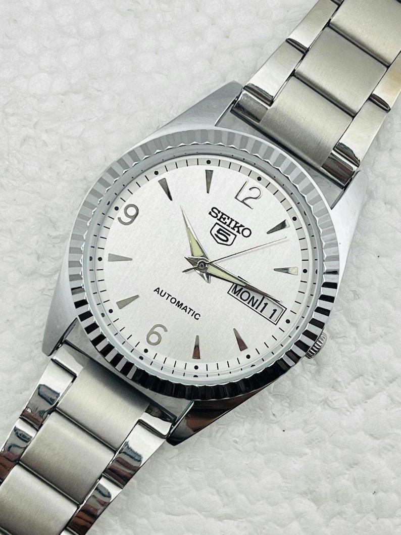 May include: Silver-toned Seiko 5 automatic watch with a white dial. The watch features black hour markers, a day-date display, and a fluted bezel. The watch band is made of silver-toned metal.