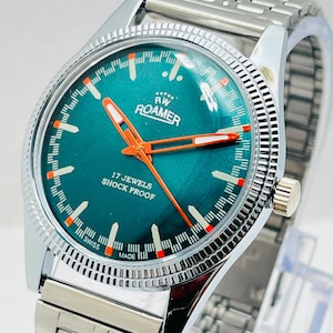 Può includere: Un orologio da polso analogico color argento con quadrante verde acqua, lancette arancioni e bianche, indici delle ore arancioni e bianchi e un cinturino a maglie in metallo. Il quadrante riporta il testo ROAMER, 17 JEWELS, SHOCK PROOF e SWISS MADE.