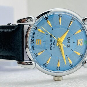 Op de afbeelding: Een zilverkleurig Oris horloge met een lichtblauwe wijzerplaat en gouden wijzers en markeringen. Het horloge heeft een zwarte leren band. De wijzerplaat toont "SWISS MADE", "ORIS" en "JEWELS SHOCK PROOF". De diameter is ongeveer 3,5 cm.