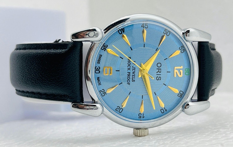 Op de afbeelding: Een zilverkleurig Oris horloge met een lichtblauwe wijzerplaat en gouden wijzers en markeringen. Het horloge heeft een zwarte leren band. De wijzerplaat toont "SWISS MADE", "ORIS" en "JEWELS SHOCK PROOF". De diameter is ongeveer 3,5 cm.