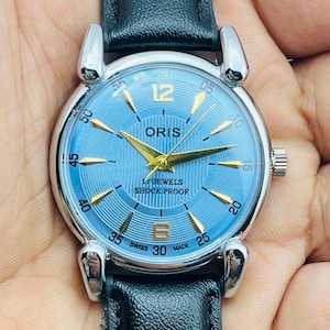 Op de afbeelding: Een polshorloge met een lichtblauwe wijzerplaat en goudkleurige wijzers en markeringen. Het horloge heeft een zilverkleurige kast en een zwarte leren band. De wijzerplaat toont "ORIS", "17 JEWELS" en "SHOCK PROOF".
