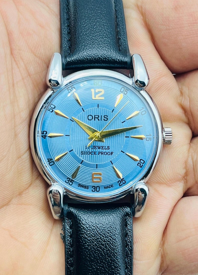 Op de afbeelding: Een polshorloge met een lichtblauwe wijzerplaat en goudkleurige wijzers en markeringen. Het horloge heeft een zilverkleurige kast en een zwarte leren band. De wijzerplaat toont "ORIS", "17 JEWELS" en "SHOCK PROOF".