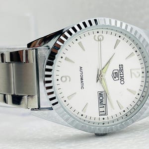 May include: A silver-toned Seiko 5 automatic wristwatch with a white dial. The watch features a fluted bezel, a date display, and a stainless steel band. The dial displays the day and date.