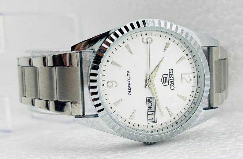 May include: A silver-toned Seiko 5 automatic wristwatch with a white dial. The watch features a fluted bezel, a date display, and a stainless steel band. The dial displays the day and date.