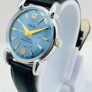 Op de afbeelding: Een zilverkleurig Oris horloge met een lichtblauwe wijzerplaat en gouden wijzers. Het horloge heeft een zwarte leren band en de tekst "17 JEWELS SHOCK PROOF" is zichtbaar op de wijzerplaat. Het horloge wordt op een witte standaard getoond.