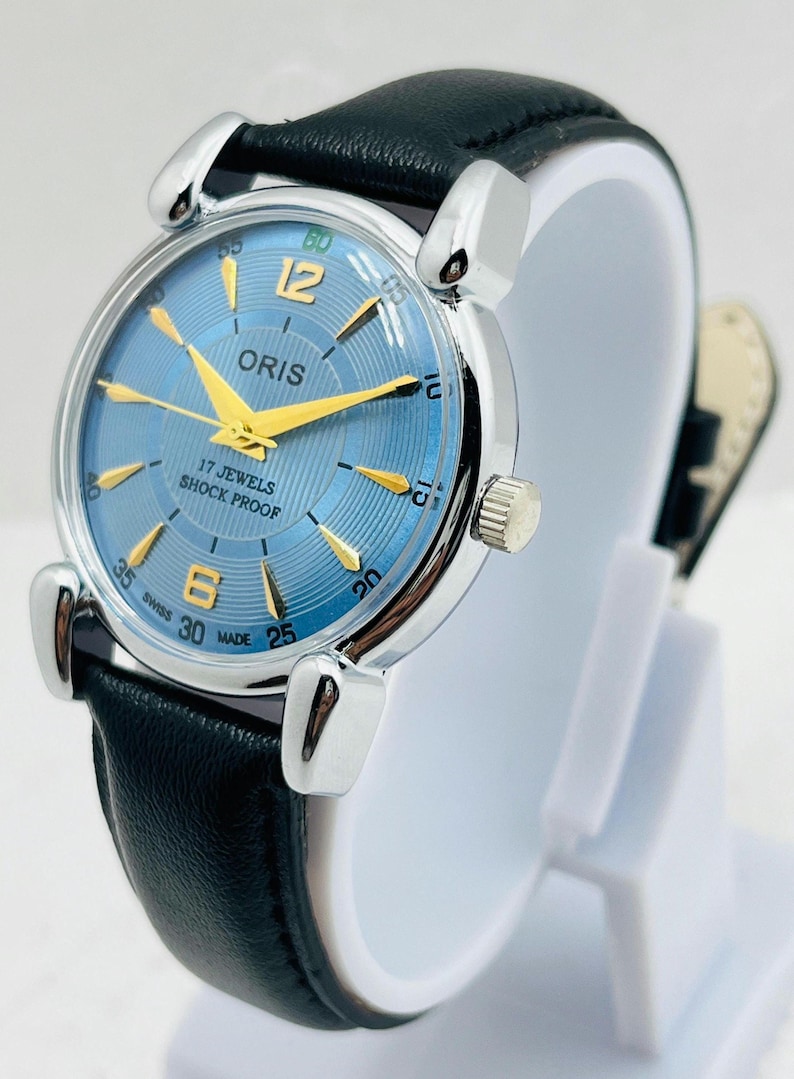 Op de afbeelding: Een zilverkleurig Oris horloge met een lichtblauwe wijzerplaat en gouden wijzers. Het horloge heeft een zwarte leren band en de tekst "17 JEWELS SHOCK PROOF" is zichtbaar op de wijzerplaat. Het horloge wordt op een witte standaard getoond.