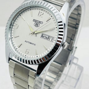 May include: A silver Seiko 5 automatic watch with a white dial. The watch features a fluted bezel, a stainless steel band, and a day-date display. The watch face displays the day as "MON 11".