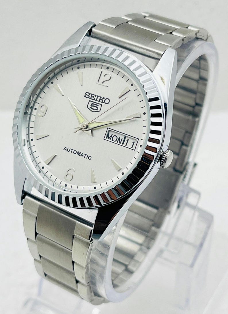 May include: A silver Seiko 5 automatic watch with a white dial. The watch features a fluted bezel, a stainless steel band, and a day-date display. The watch face displays the day as "MON 11".