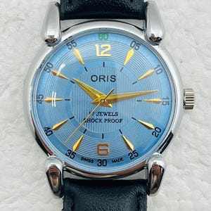 Op de afbeelding: Een polshorloge met een lichtblauwe wijzerplaat, gouden wijzers en een zwarte leren band. De wijzerplaat toont de merknaam "ORIS" en de tekst "7 JEWELS SHOCK PROOF". Het horloge heeft een zilverkleurige kast en een zwarte leren band.