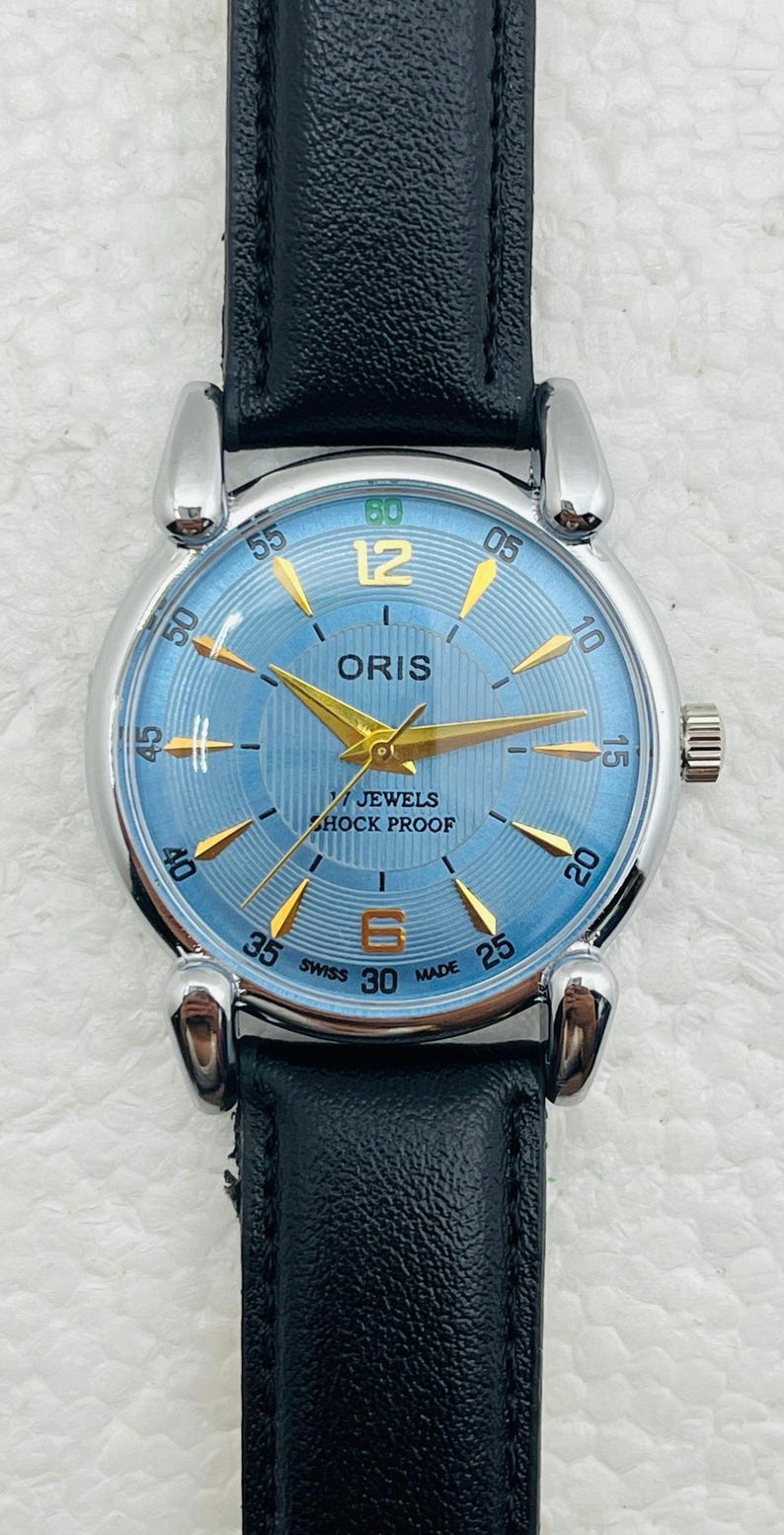 Op de afbeelding: Een polshorloge met een lichtblauwe wijzerplaat, gouden wijzers en een zwarte leren band. De wijzerplaat toont de merknaam "ORIS" en de tekst "7 JEWELS SHOCK PROOF". Het horloge heeft een zilverkleurige kast en een zwarte leren band.