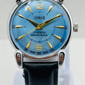 Op de afbeelding: Een vintage ORIS polshorloge met een lichtblauwe wijzerplaat en goudkleurige wijzers en markeringen. Het horloge heeft een zwarte leren band en een zilverkleurige roestvrijstalen kast. De wijzerplaat toont "17 JEWELS SHOCK PROOF" en "SWISS MADE".