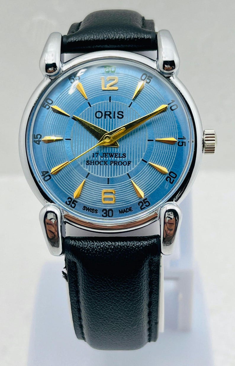 Op de afbeelding: Een vintage ORIS polshorloge met een lichtblauwe wijzerplaat en goudkleurige wijzers en markeringen. Het horloge heeft een zwarte leren band en een zilverkleurige roestvrijstalen kast. De wijzerplaat toont "17 JEWELS SHOCK PROOF" en "SWISS MADE".