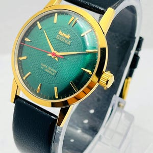 May include: A gold wristwatch with a green face and a black leather strap. The watch has gold hands and markers, with the text "Sona" and "Para Shock 17 Jewels". The watch is displayed on a clear stand.