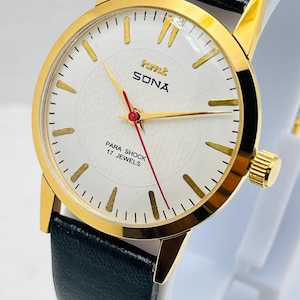 May include: A gold wristwatch with a white face, gold hands, and a red second hand. The watch has a black leather strap and the face reads "SONA" and "PARA SHOCK 17 JEWELS".