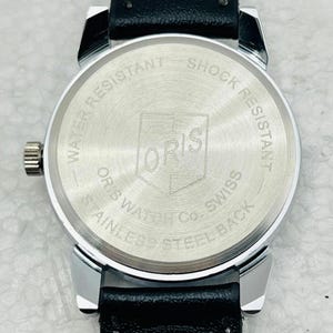 Op de afbeelding: Close-up van de achterkant van een zilverkleurig horloge met een zwarte leren band. De wijzerplaat heeft de merknaam "ORIS" en de tekst "WATER RESISTANT SHOCK RESISTANT STAINLESS STEEL BACK".