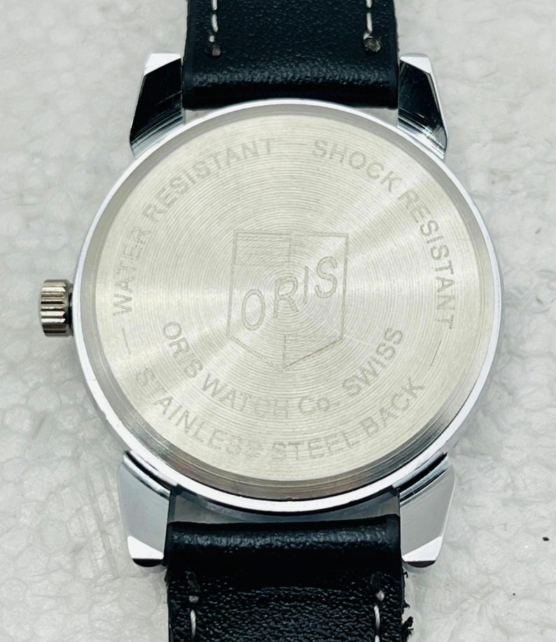 Op de afbeelding: Close-up van de achterkant van een zilverkleurig horloge met een zwarte leren band. De wijzerplaat heeft de merknaam "ORIS" en de tekst "WATER RESISTANT SHOCK RESISTANT STAINLESS STEEL BACK".