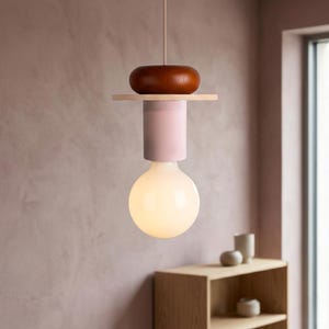 May include: A modern pendant light fixture with a large, round, illuminated bulb. The fixture features a wooden top, a light pink cylinder, and a wooden accent. The light hangs from a thin cord, showcasing a minimalist design.